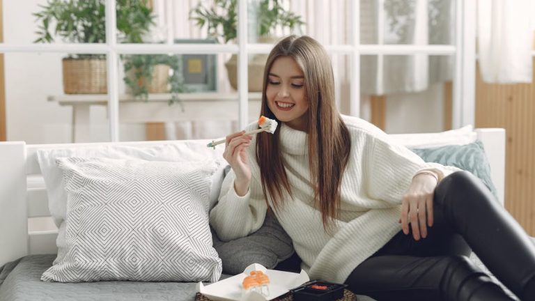 Smiling young lady in warm knitted sweater enjoying fresh sushi on wicker tray while relaxing on cozy sofa with soft cushions in modern living room